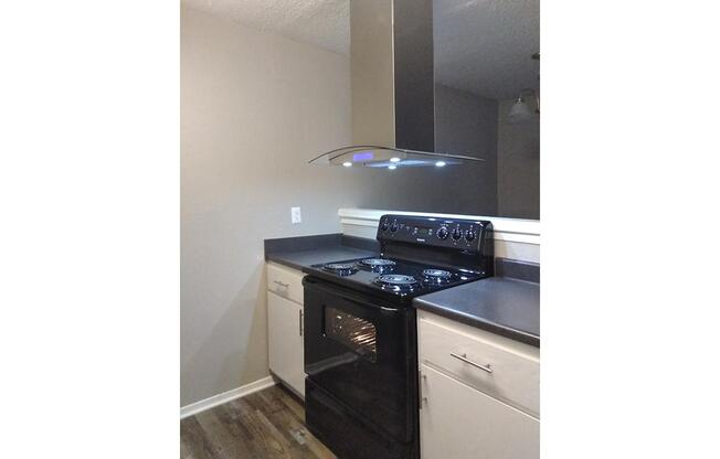 A modern kitchen featuring a black freestanding oven and stove with four burners. There is a sleek stainless steel range hood above the stove equipped with under-cabinet lighting. The cabinetry is a mix of light and dark finishes on a wooden floor, providing a contemporary look.