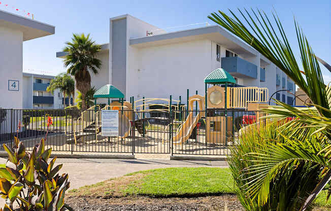 A playground with a slide and a green canopy is surrounded by a fence and a building with a number 4 on it.