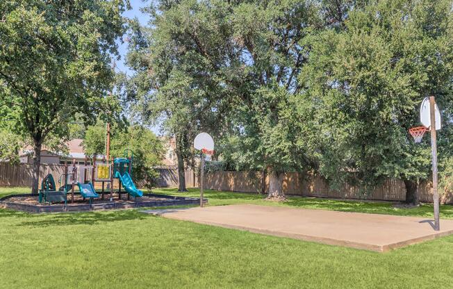 A sunny playground featuring a blue slide and climbing structure, adjacent to a basketball court with two hoops. The area is surrounded by tall green trees and a wooden fence, with well-maintained grass covering the ground.