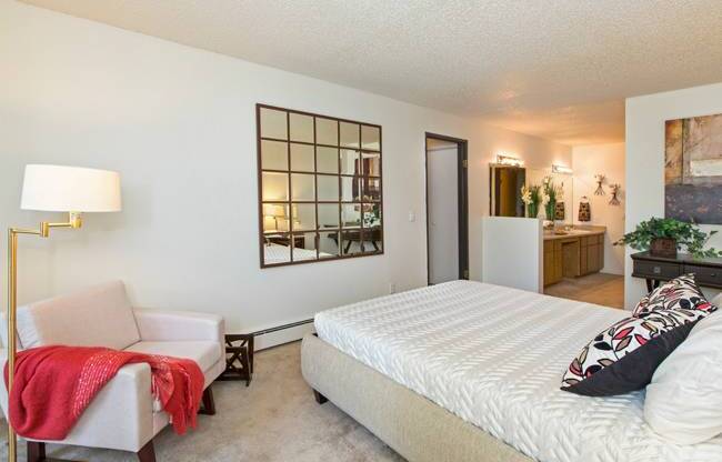A bright bedroom here at Eagle Ridge showcasing a spacious sleeping area with a neatly made bed, plush carpeted flooring, a cozy upholstered accent chair with a floor lamp, decorative wall mirror, and an open layout that leads toward a vanity and bathroom area, creating a comfortable and functional retreat with warm lighting and clean finishes.