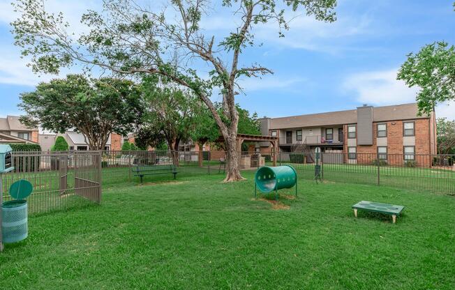A spacious green dog park enclosed by a fence, featuring a few benches, trees, and play equipment, including agility tunnels. In the background, there are residential buildings. The sky is clear, adding to the inviting atmosphere of the park.