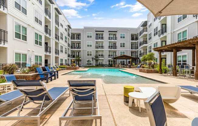 A pool surrounded by chairs and umbrellas in front of apartment buildings.