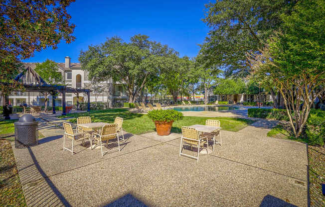 Outdoor lounge area at Saxony at Chase Oaks Apartments in Dallas, TX, featuring a shaded pergola with seating, patio tables with chairs, a lush green lawn, and a sparkling swimming pool surrounded by mature trees and apartment buildings.