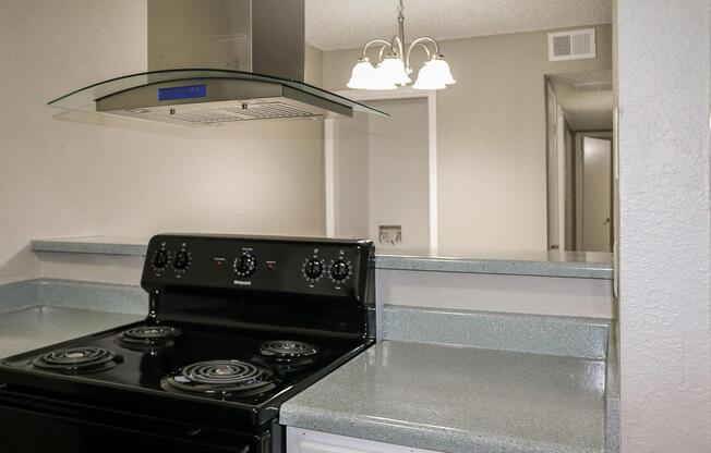 A modern kitchen featuring a black stove with four burners, a sleek silver hood above, and a stone countertop. In the background, there is a hint of an open area with a light-colored wall and a doorway seen in the distance. The kitchen has a clean and contemporary appearance.