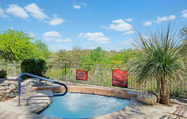 A relaxing outdoor spa here at Mirador featuring a circular hot tub with tiled surround and curved stainless handrail, set against desert landscaping and scenic hillside views. Wrought iron fencing, natural stone accents, and nearby lounge seating create a peaceful retreat ideal for unwinding under sunny Arizona skies.