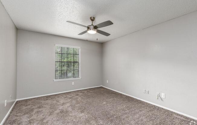 Empty room with light gray walls and plush brown carpet. A simple ceiling fan is mounted above, and there is a window with blinds allowing natural light to enter. The room appears bright and spacious, ideal for personalization or furnishing. Electrical outlets are visible on the wall.