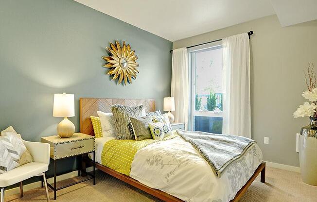 A bedroom with a bed, nightstand, chair, and a sunburst mirror on the wall. at Kirkland Crossing Apartments, Washington, 98033