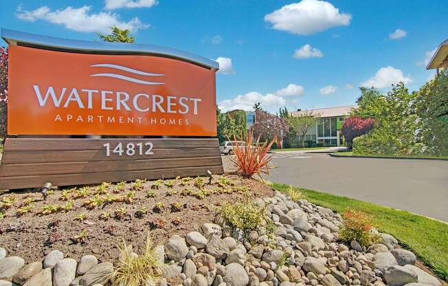 An inviting community entrance sign here at Watercrest featuring a bold orange monument sign, landscaped rock garden with greenery, and a clean driveway leading toward attractive residential buildings under a bright blue sky.