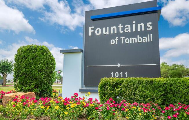 Sign for "Fountains of Tomball" with the address 1011, surrounded by colorful flowers and manicured bushes under a partly cloudy sky.