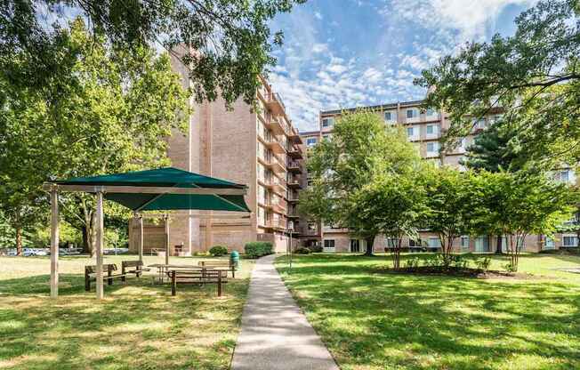 A green canopy pavilion is situated in a grassy area with a paved walkway leading to apartment buildings.