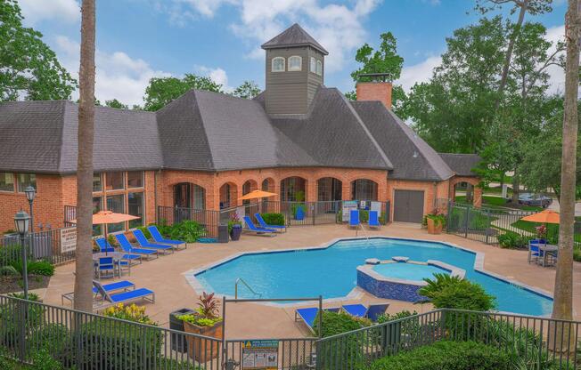 Aerial view of a resort-style pool area featuring a large blue swimming pool, a hot tub, lounge chairs, and umbrellas. Surrounding the pool are well-maintained shrubs and trees, with a clubhouse in the background showcasing a modern architectural design. Clear skies and a lush landscape enhance the inviting atmosphere.