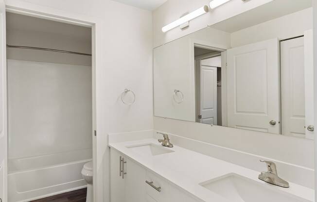 A full bathroom with double vanity sinks, white countertops and cabinets, a large mirror, and a door to the toilet and shower.
