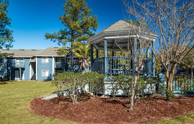A gazebo is in the middle of a grassy area with a tree and shrubs around it.
