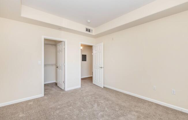 A spacious room featuring light-colored walls and carpet flooring. Two open doors lead to a closet and an adjoining room. The ceiling has a slight recess, contributing to the overall bright and airy feel of the space.