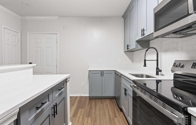 Modern kitchen featuring light blue and gray cabinets, a stainless steel stove, and a sleek countertop. The space is well-lit with light-colored walls and laminate flooring, creating a clean and inviting atmosphere. A door is visible, leading to another room.
