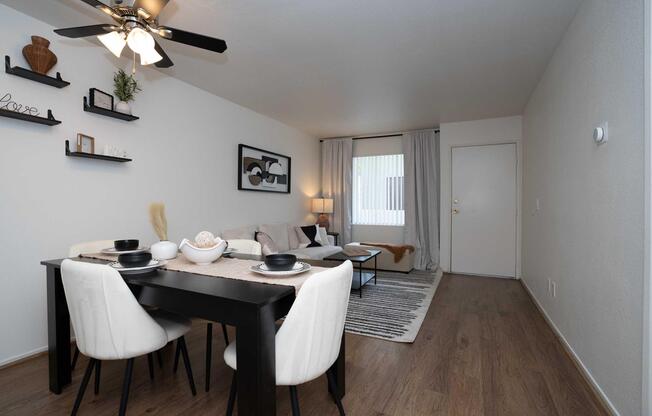 A modern living space features a dining table set for four with black and white tableware. The room has a stylish sofa, neutral decor, and a ceiling fan. A large window with light curtains allows natural light. Decorative shelves and a cozy area rug complete the inviting atmosphere.