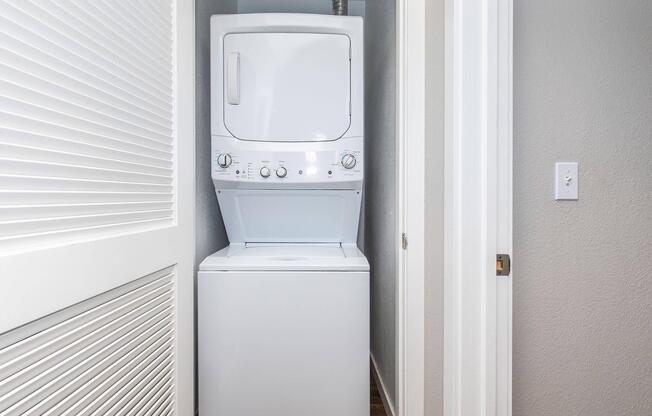 A stacked washer and dryer unit located in a narrow laundry space with light gray walls and a white door partially closed, revealing modern appliances designed for space-saving in a home.