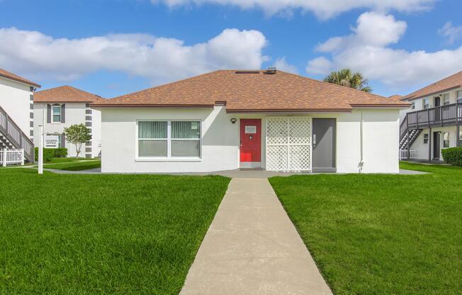 A single-story white house with a red door, featuring a latticework screen on one side. The house is surrounded by lush green grass and has a clear blue sky with fluffy white clouds in the background. There are two multi-unit buildings in the background with balconies, creating a pleasant residential setting.