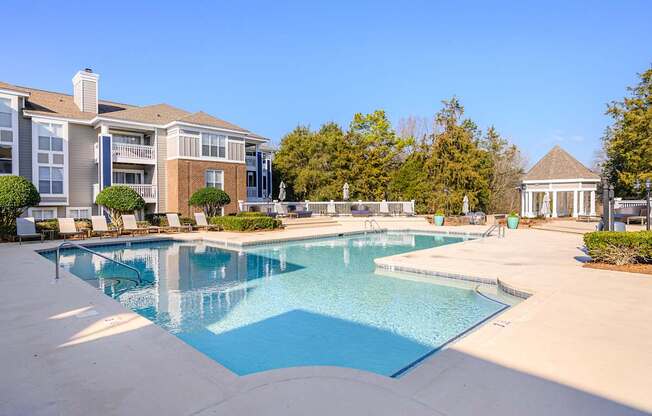 A resort-style swimming pool here at Retreat at McAlpine Creek featuring a spacious pool with clear blue water, wide deck areas, and multiple lounge chairs for relaxing. Surrounding apartment buildings, manicured landscaping, and a nearby gazebo create a peaceful outdoor setting ideal for unwinding or spending time with neighbors.