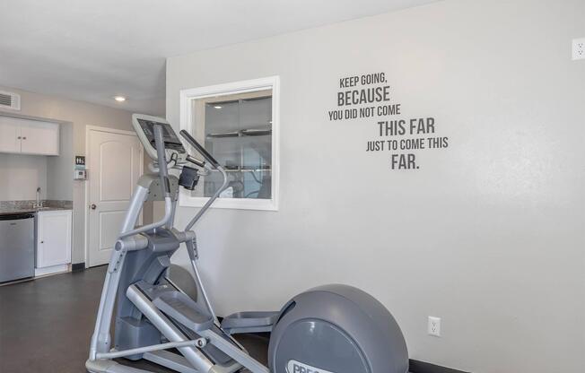 A modern gym interior featuring an elliptical machine beside a wall with inspirational text that reads, "Keep going, because you did not come this far just to come this far." The space has a bright, inviting atmosphere with neutral-colored walls.