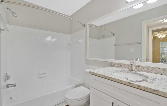 A clean and modern bathroom featuring a white bathtub with a shower, a large mirror above a granite countertop sink, and a toilet. The walls are painted in a light color, creating a spacious feel, and there is a glimpse of a light fixture and window in the background.