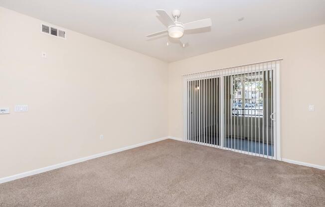 Spacious, empty room with light beige walls and plush carpet. A ceiling fan is mounted above, and there is a sliding glass door with vertical blinds leading to an outdoor area. The room is bright and inviting, suitable for various furnishings.
