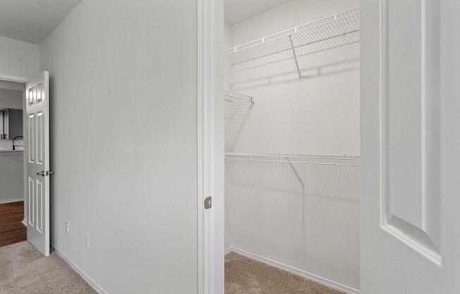 A view of a partially open closet with empty wire shelving, featuring light-colored walls and carpeted flooring. The doorway to the closet is in focus, with a neutral color scheme creating a spacious feel. In the background, a glimpse of a kitchen area can be seen.