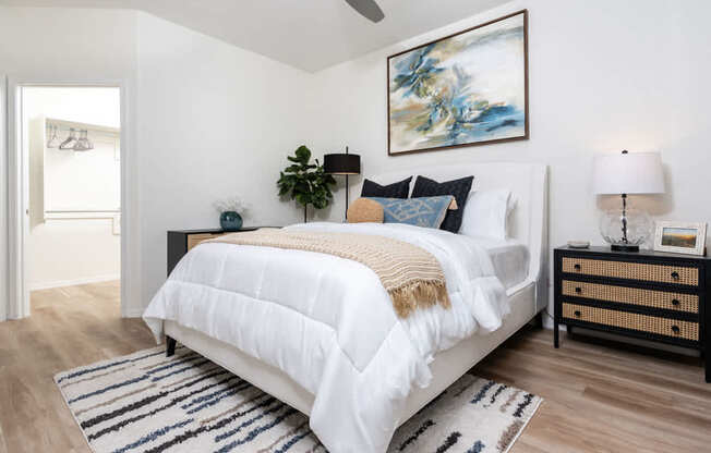 A bedroom with a large bed, a painting on the wall, and a wooden nightstand.