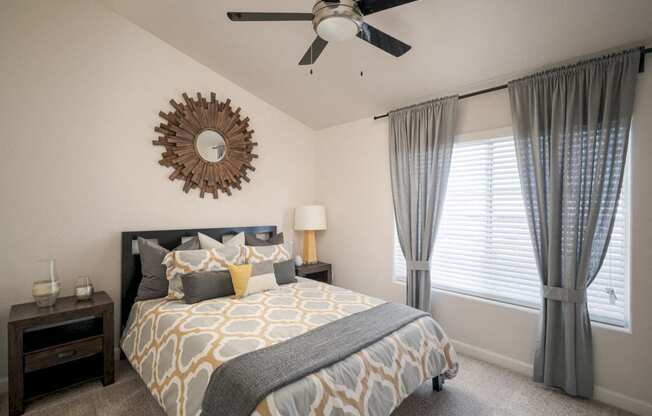 Model bedroom with wall to wall carpet, ceiling fans, queen bed, and large windows