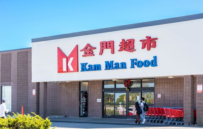 A storefront of a Kam Man Food store with people walking by.