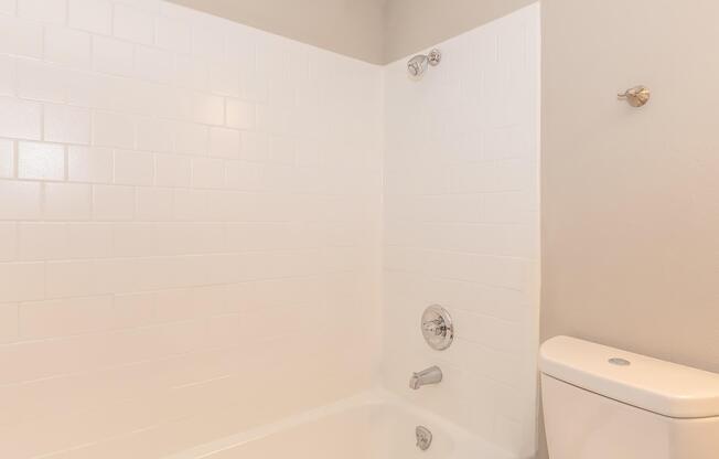 A clean, modern bathroom featuring a white tiled bathtub with a shower head, a chrome faucet, and a white toilet. The walls are a light gray color, and there is a simple design with no clutter, creating a spacious and inviting atmosphere.