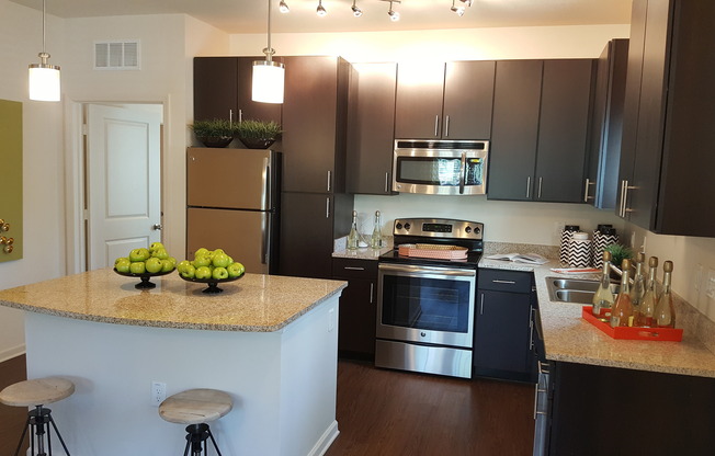 Photo of kitchen with large island, granite counter-tops, modern cabinetry, microwave, electric stove, dishwasher, refrigerator, and double sided sink, from a different angle, showing seating at the island and both pendant and track lightin