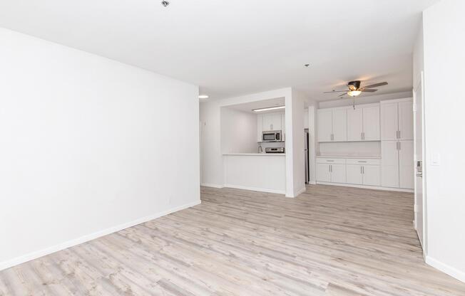 Spacious, modern living area featuring light wood flooring and neutral-colored walls. The far end shows an open kitchen with white cabinetry, stainless steel appliances, and a ceiling fan. Natural light enhances the bright and airy atmosphere, creating a welcoming environment.