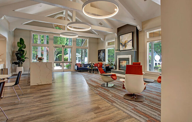 A bright resident lounge here at Portsmith showcasing vaulted ceilings with exposed beams, modern circular pendant lighting, and expansive windows that bring in natural light and leafy outdoor views, with contemporary lounge chairs in warm accent tones, a statement fireplace with artwork, wood-style flooring, and an open kitchen island that creates a welcoming space for relaxing or gathering.