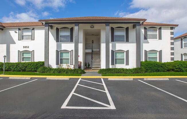A two-story apartment building with a white exterior and dark gray accents. There are stairs leading to the entrance, flanked by large windows. The surrounding landscape features green shrubbery and a clear blue sky. Parking spaces are visible in front of the building, with a few marked lines.