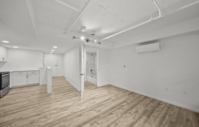 A spacious, modern apartment interior featuring light-colored walls and wood-style flooring. The open layout includes a kitchen area with white cabinets and appliances, a bright living space, and a bathroom visible through an open door. Natural light fills the room, creating an inviting atmosphere.