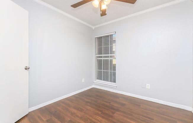 A simple, empty room with light gray walls and hardwood flooring. It features a ceiling fan with light fixtures, a single white door on the left, and a window with white blinds allowing natural light. The space feels bright and open, perfect for various uses.