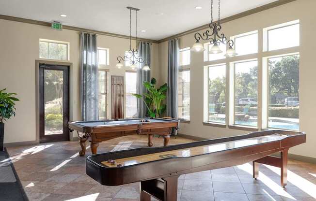A game room here at Landing at Mansfield with a wooden pool table and shuffleboard table, tall bright windows, dark metal chandeliers, and warm tile flooring overlooking the pool area.
