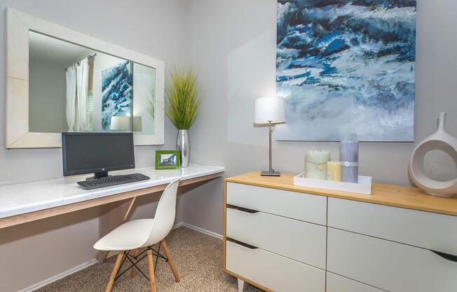 A cozy home office setup featuring a modern desk with a computer, a stylish chair, and a decorative lamp. The wall displays a large abstract painting in blue tones, and a potted plant adds a touch of greenery. A dresser with a minimalist design sits adjacent to the desk, enhancing the space's contemporary feel.