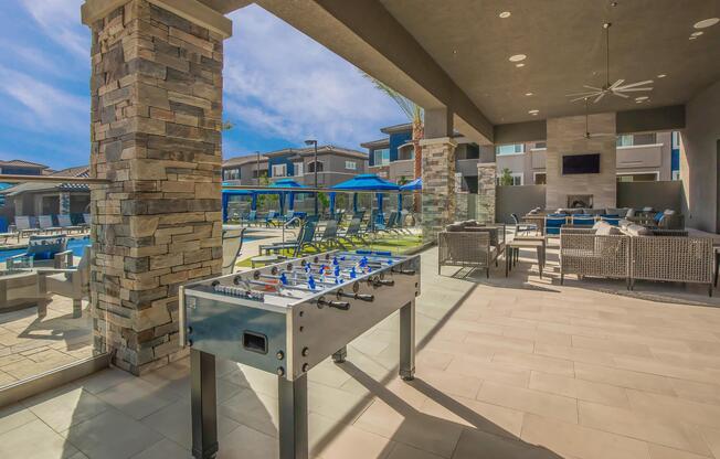 A modern outdoor lounge area featuring a foosball table in the foreground. Behind it are comfortable seating arrangements and a swimming pool area with blue umbrellas. The space is well-lit and has stone accents, providing a relaxing atmosphere.