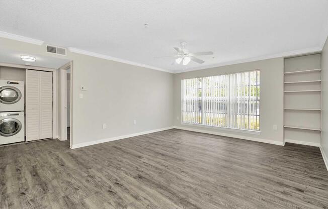 Spacious living area with light-colored walls and hardwood flooring. Features include a ceiling fan, large windows with blinds, and a built-in shelf. A laundry area is visible through an open door on the left, adding convenience to the space. The room is well-lit and inviting.