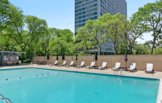 A swimming pool surrounded by trees and chairs with a building in the background.