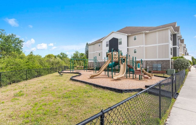 A playground with a slide and a green fence in front of a building.