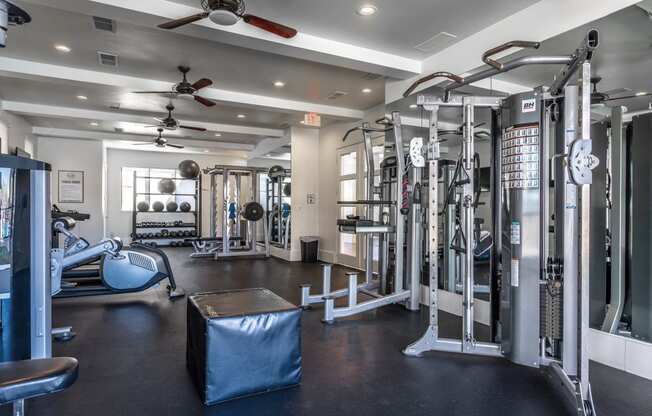 Studio Apartments in Rowlett, TX - Village of Rowlett - Fitness Center with Weights, Strength Training Machines, Ceiling Fans, Large Mirrors, and Windows.