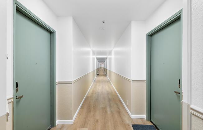 A long, well-lit hallway with two green doors on either side. The walls are painted white, and there is a wooden floor. Decorative mats are placed in front of the doors, leading down the straight corridor toward a distant perspective.