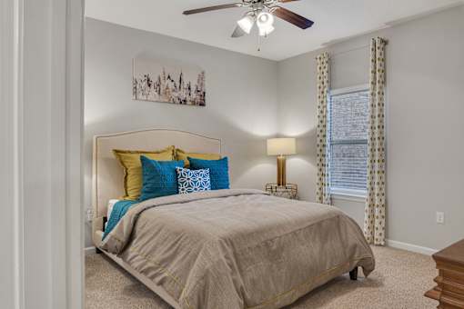 A bedroom with a bed, pillows, a ceiling fan, and a painting on the wall.