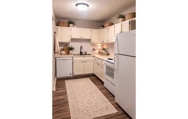 A modern kitchen showcasing white cabinetry, a countertop with a sink, and essential appliances including a refrigerator and an oven. The space is decorated with green plants and decorative baskets, complemented by a light decorative rug on the floor, creating a warm and inviting atmosphere.