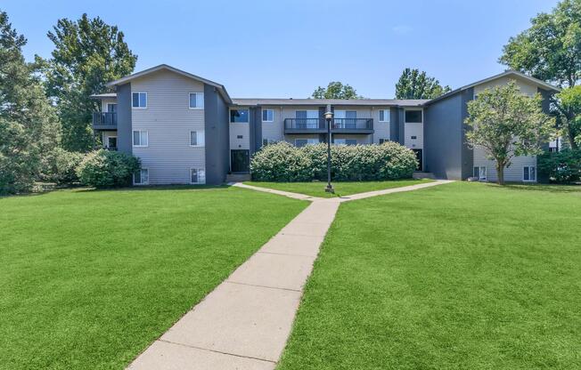 A well-maintained apartment complex with three buildings surrounded by green grass and shrubs. A paved pathway leads through the lawn, connecting the buildings. Trees are visible in the background under a clear blue sky. Ideal for showcasing residential living spaces.