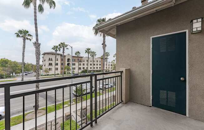 A balcony with a green door and a black railing overlooks a street with palm trees.