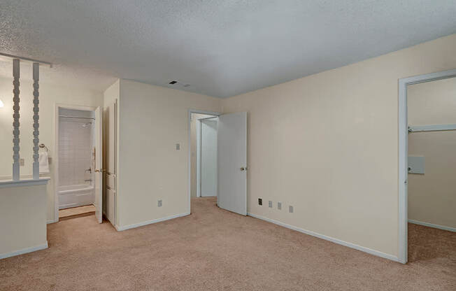A spacious neutral bedroom here at Summertree Place featuring plush carpet flooring, soft beige walls, a textured ceiling, an open layout with multiple doorways, access to a connected bathroom, and a walk-in closet area, offering flexibility and comfort within a bright, functional space.